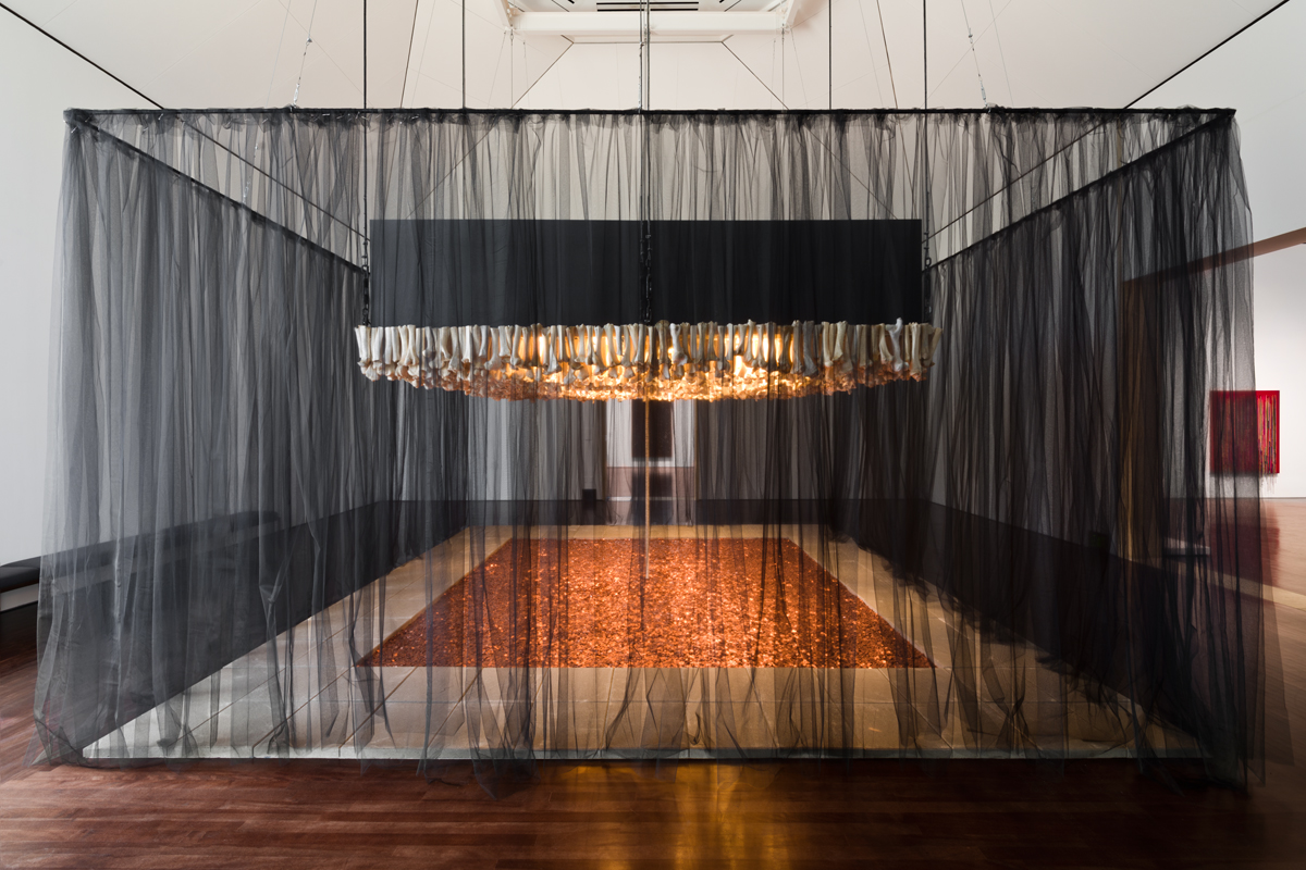 Photo of Cildo Meireles' Missão/Missões [Mission/Missions] (How to Build Cathedrals) , 1987. The piece is made up of 600,000 coins, 800 communion wafers, 2,000 cattle bones, 80 paving stones, and black cloth. The paving stones are positioned into a rectangle with the interior being full of pennies. In the middle of the pit of pennies is a stack of communion wafers that go towards the top of the piece. The top of the piece is a "roof" of cattle bones positioned vertically. The entire piece is surrounded by a black cloth.