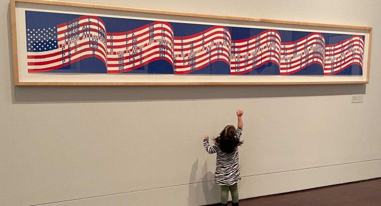 A toddler girl looking up and pointing at a long horizontal print of the USA flag hanging on the wall.