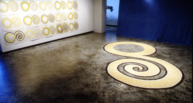 Installation view of artworks, a swirling pattern on the floor, repeated on the wall in a pattern-like presentation.